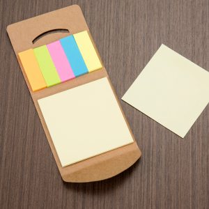 Bloco de anotações em papel reciclado com cor da capa marrom e por dentro bloco de anotação pequeno retangular na cor (laranja, verde, rosa, azul, e amarelo) seguindo da esquerda para a direita e bloco de anotação quadrado na cor beje e uma folha quadrada na cor beje separada do kit em cima de uma mesa de madeira marrom.