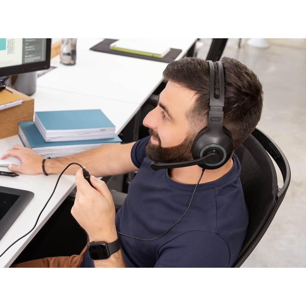 Foto de uma um home branco de barba de camiseta azul marinho, relogio preto no pulso esquerdo, calça na cor beje, e usando um fone de ouvido com microfone preto com cabo também preto, e segurando na mão esquerda o controle de volume do fone que fica no cabo do mesmo Sentado em uma cadeira preta olhando para um computador segurando um mouse, sobre uma mesa de cor branca com livros de capa azul, a ponta do lado direito de uma monitor de computador de mesa (desktop).